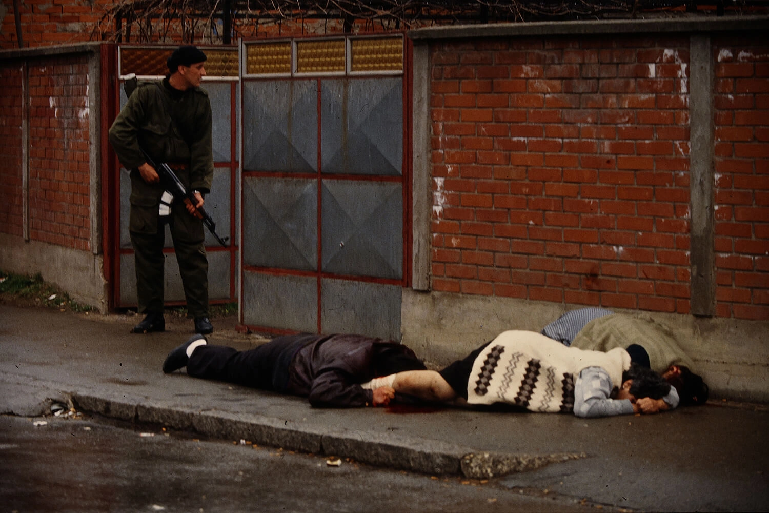 Civilians lie on the ground after they were gunned down in Bijeljina, on April 2, 1992. An armed member of Arkan's Tigers walks behind them.
